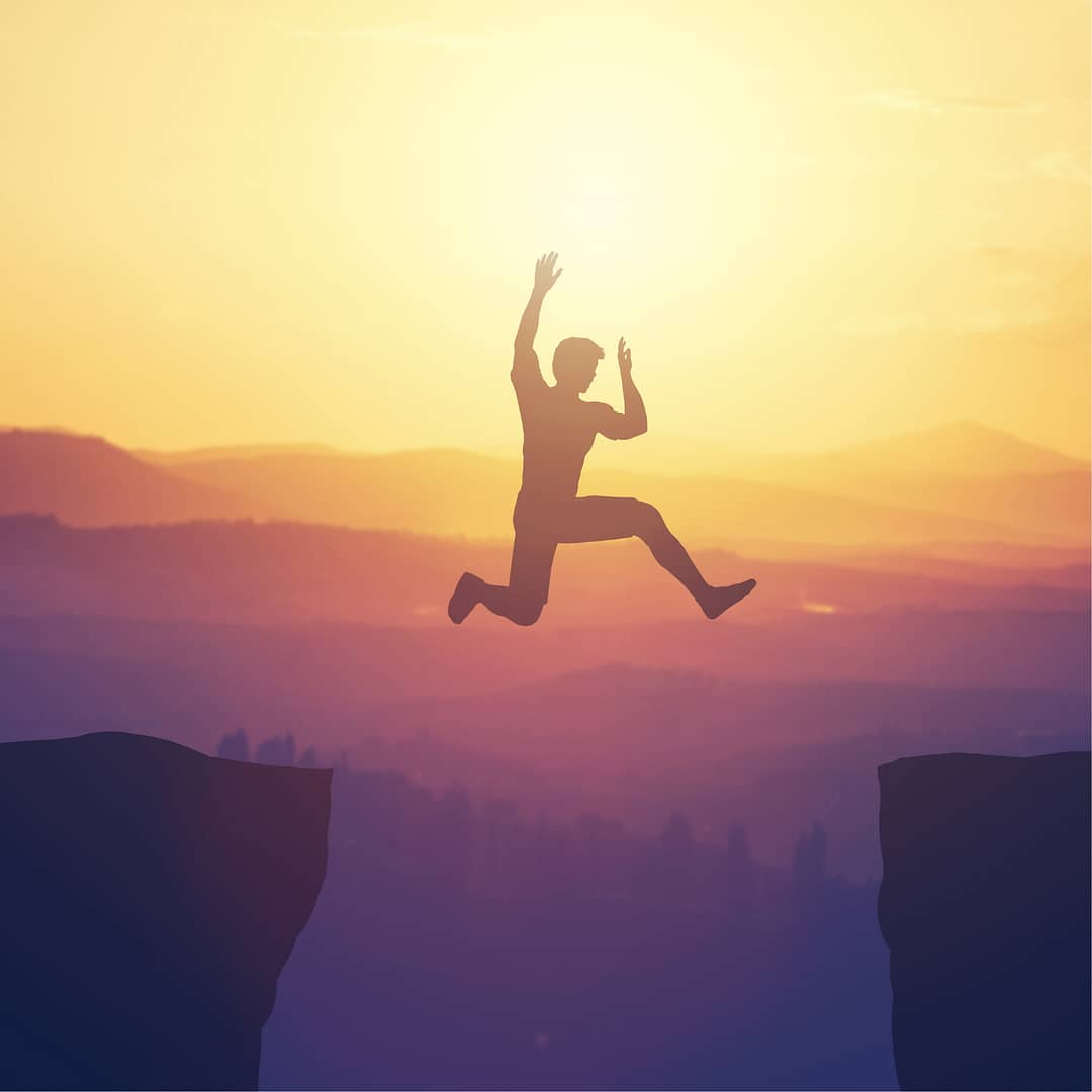 A human being jumping across a gorge between two cliffs with the sun in the background.